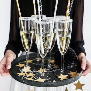 May include: A tray of champagne flutes with gold and black striped straws. The tray is decorated with gold star confetti and a gold star garland.