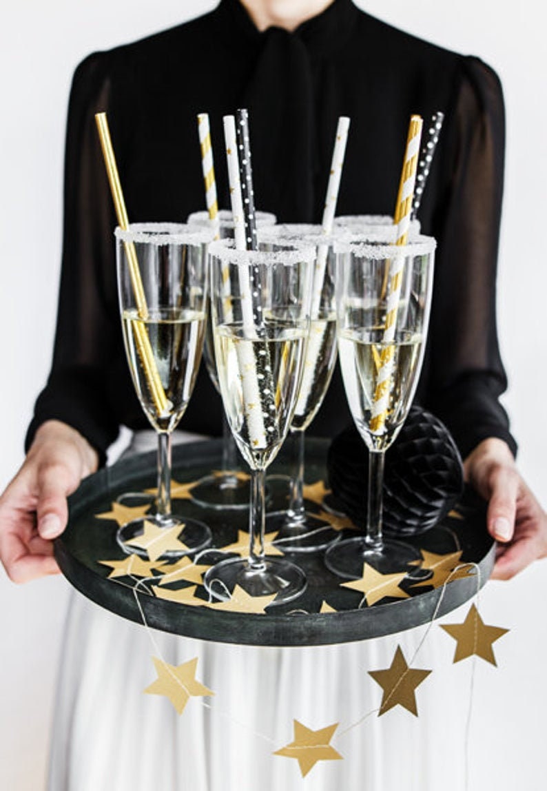 May include: A tray of champagne flutes with gold and black striped straws. The tray is decorated with gold star confetti and a gold star garland.