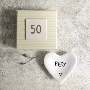 Op de afbeelding: Een vierkante, crèmekleurige geschenkdoos met een kleine, witte vierkant met het grijze getal "50". Een witte, hartvormige schaal met het woord "Fifty" en een klein hartsymbool. Een takje gedroogd plantmateriaal ligt naast de schaal.