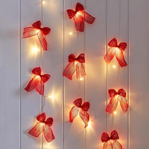 May include: A decorative string of red ribbon bows with warm white fairy lights. The bows are arranged vertically against a white wooden backdrop. The lights are evenly spaced, creating a festive and decorative display.
