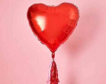 Puede incluir: Un gran globo rojo brillante en forma de corazón con una borla. El globo está inflado y parece estar hecho de un material metálico. La borla es una mezcla de rosa y rojo. El fondo es de color rosa liso.