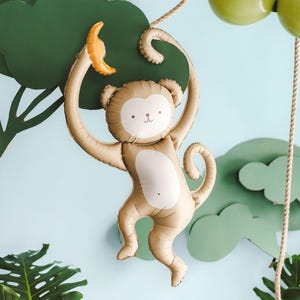 May include: A tan monkey-shaped foil balloon featuring a cream-colored face and belly, with a small yellow banana held in one raised arm. The monkey has a curled tail and a simple printed facial expression with two eyes, a nose, and a mouth.
