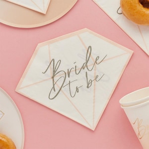 May include: A diamond-shaped napkin with the words "Bride to be" in elegant script. The napkin has a white center with a pale pink border and accents. Other party supplies, including a plate, cup, and donut, are visible on a pink background.