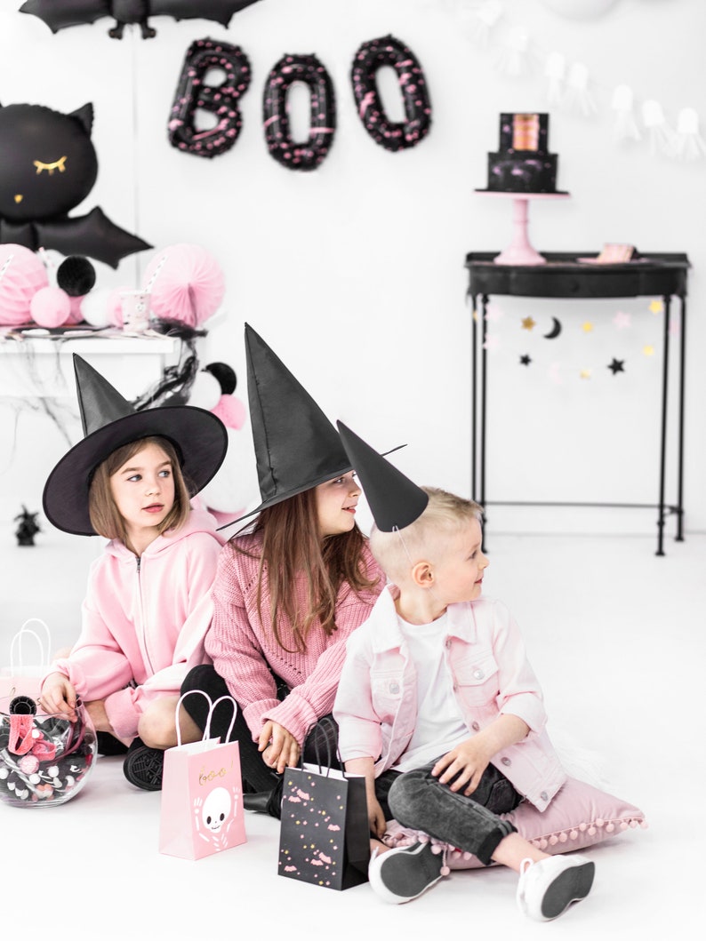 K&ouml;nnte beinhalten: Drei Kinder in rosa und schwarzen Halloween-Kost&uuml;men, darunter Hexenh&uuml;te, halten rosa und schwarze Geschenkt&uuml;ten. Der Hintergrund zeigt schwarze und rosa Halloween-Dekorationen, darunter eine Fledermaus, ein BOO-Schild und eine schwarze und rosa Torte.
