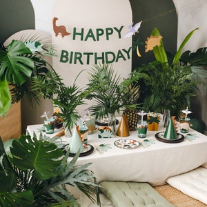 May include: A festive dinosaur-themed birthday party setup. The table is decorated with plates, cups, and party hats. A banner reads "HAPPY BIRTHDAY" with dinosaur cutouts. Lush greenery and woven lampshades complete the scene.