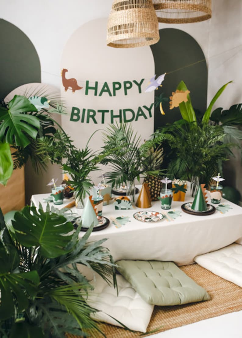 May include: A festive dinosaur-themed birthday party setup. The table is decorated with plates, cups, and party hats. A banner reads "HAPPY BIRTHDAY" with dinosaur cutouts. Lush greenery and woven lampshades complete the scene.