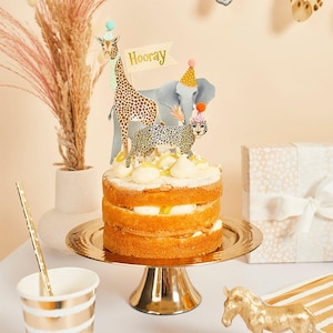 May include: A layered cake with white frosting and animal-themed cake toppers, including a giraffe, elephant, and leopard, all wearing party hats. A banner that says "Hooray" is also on the cake. The cake sits on a gold cake stand.