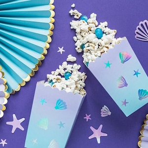 May include: Two purple and blue popcorn boxes with a mermaid theme. The boxes are decorated with iridescent seashells and stars. The boxes are filled with white popcorn and blue candy.