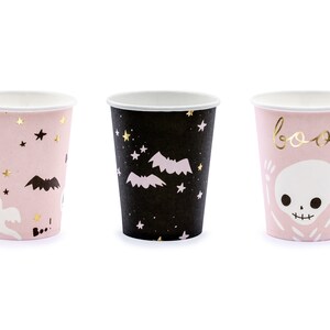 May include: Three paper cups with Halloween-themed designs. One cup is black with bats and stars, another is pink with a ghost, pumpkin, and stars, and the third is pink with a skeleton and the word "boo!"