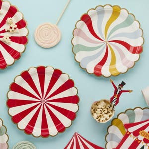 May include: Overhead shot of circus-themed party supplies on a light blue surface. Includes red and white striped plates, plates with colourful swirl designs, popcorn, and a lollipop. Gold scalloped edges.