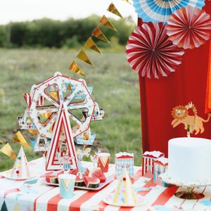 May include: A red and white striped tablecloth with a white paper Ferris wheel, red and white striped paper cups, and a white cake with a lion topper. There are also red and white striped paper boxes and a plate of watermelon slices on the table.