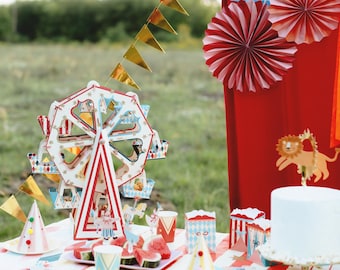 May include: A red and white striped tablecloth with a white paper Ferris wheel, red and white striped paper cups, and a white cake with a lion topper. There are also red and white striped paper boxes and a plate of watermelon slices on the table.
