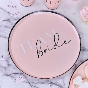 May include: Pink paper party plate with black trim and the text "TEAM bride" in white and black lettering.
