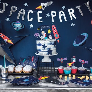 May include: A space-themed party table setting with a blue and white cake decorated with astronaut figurines, cupcakes with rocket ship toppers, and a variety of space-themed decorations, including stars, planets, and rockets.