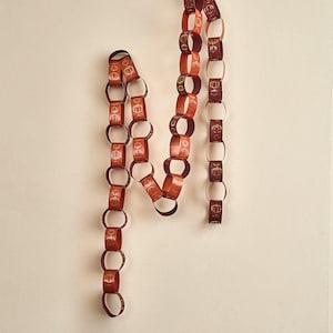 May include: A decorative paper chain garland featuring interlocking loops in shades of orange, dark red, and brown. Each individual paper link is printed with a repeating gold-colored line art illustration of a pumpkin, creating a continuous pattern.