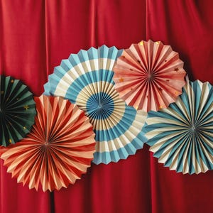 May include: Six paper fans in various colors and patterns, including red, white, blue, green, and pink. The fans are arranged in a semi-circle on a red background.