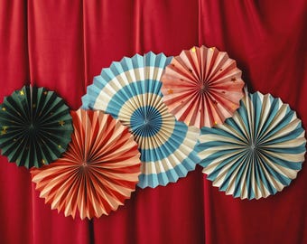 Könnte beinhalten: Sechs Papierfächer in verschiedenen Farben und Mustern, darunter Rot, Weiß, Blau, Grün und Pink. Die Fächer sind in einem Halbkreis auf einem roten Hintergrund angeordnet.