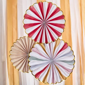 Pode incluir: Três leques decorativos de papel em vermelho e branco, castanho e branco, e cores pastel com borda dourada. Os leques estão dispostos em frente a uma cortina listrada amarela e branca, criando um ambiente festivo.