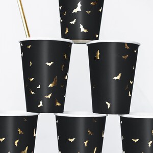 May include: Black paper cups with a white rim and gold bat silhouettes. The cups are stacked in a pyramid shape, with a gold straw visible. These cups are perfect for a Halloween or themed party.