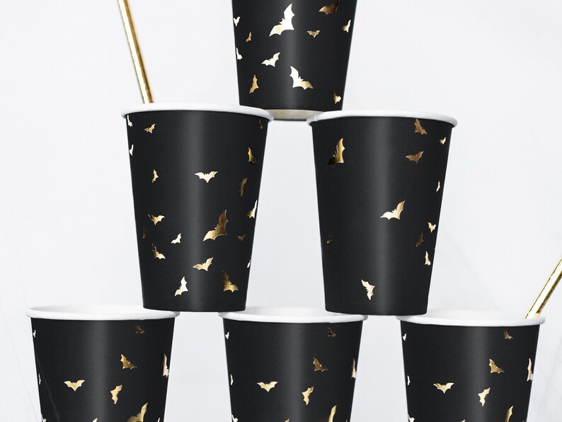 May include: Black paper cups with a white rim and gold bat silhouettes. The cups are stacked in a pyramid shape, with a gold straw visible. These cups are perfect for a Halloween or themed party.