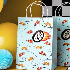 K&ouml;nnte beinhalten: Hellblaue Geschenkt&uuml;ten mit Rennwagen-Motiv. Die T&uuml;ten zeigen Illustrationen von orangefarbenen und roten Rennwagen auf einer gepunkteten Strecke. Jede T&uuml;te hat wei&szlig;e Griffe und ein dekoratives Radschild mit Flammenakzent.