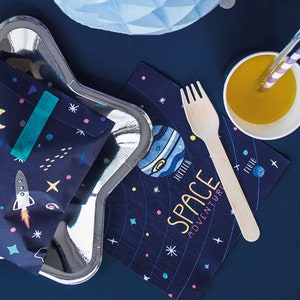 May include: A blue and white star-shaped paper plate with a blue and white space-themed paper bag inside. The bag has a teal ribbon and a rocket ship design. A blue and white space-themed napkin with the text "Space Adventure" is on the table next to a wooden fork and a white cup with orange juice.