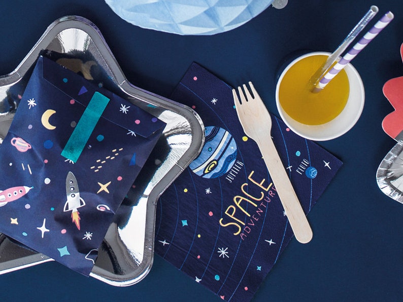 May include: A blue and white star-shaped paper plate with a blue and white space-themed paper bag inside. The bag has a teal ribbon and a rocket ship design. A blue and white space-themed napkin with the text "Space Adventure" is on the table next to a wooden fork and a white cup with orange juice.
