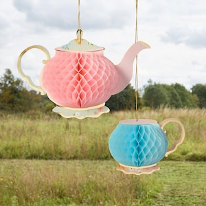 Puede incluir: Dos decoraciones colgantes de papel: una tetera rosa y una taza azul. La tetera tiene una tapa con estampado floral y detalles dorados. La taza tiene un asa y una base a juego. Ambas están suspendidas por hilos dorados.