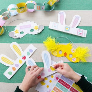 Puede incluir: Suministros para manualidades con temática de Pascua, que incluyen orejas de conejo de papel, máscaras de pollito y una cadena de papel. Las orejas de conejo y las máscaras son blancas, amarillas y de colores pastel, con pegatinas decorativas. La cadena de papel presenta ilustraciones temáticas de Pascua.
