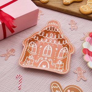 May include: A gingerbread house-shaped paper plate with white icing details, surrounded by gingerbread man and candy cane decorations. A pink gift box with a red ribbon is in the background. Festive holiday theme.