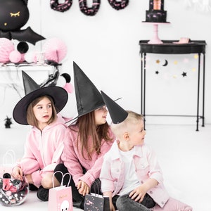 May include: Three children dressed in pink and black Halloween costumes, including witch hats, are holding pink and black gift bags. The background features black and pink Halloween decorations, including a bat, a BOO sign, and a black and pink cake.