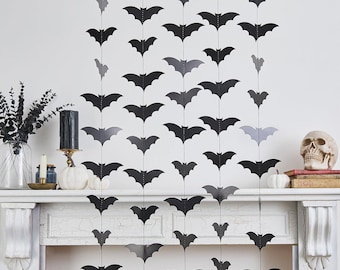 May include: Black and gray paper bat decorations hanging in rows against a white wall. The Halloween-themed decor includes a skull, pumpkins, and candles on a mantelpiece, creating a spooky atmosphere.