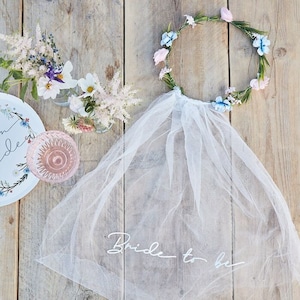 May include: White tulle veil with the words "Bride to be" written on it. The veil is paired with a floral crown with blue and pink flowers.