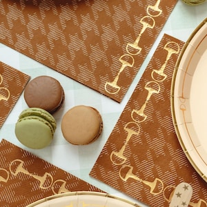 May include: An overhead shot features brown napkins with a white houndstooth pattern and gold horse bit accents. Several macarons in shades of brown and green are scattered around the napkins. A beige plate with a gold rim is also visible.
