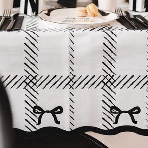 Puede incluir: Mantel blanco con un patrón de cuadros negros y detalles de lazo negro. La mesa está puesta con cubiertos negros, un plato con la inscripción "Happy Birthday" y dos macarons. El conjunto es elegante y con estilo.
