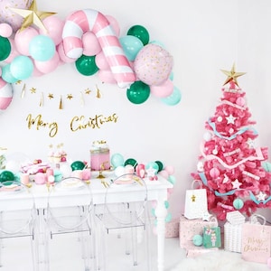 May include: A pink Christmas tree decorated with white and blue ribbons and ornaments. The tree is surrounded by white gift bags with gold stars and a sign that says "Merry Christmas". There are also pink and green balloons in the background.