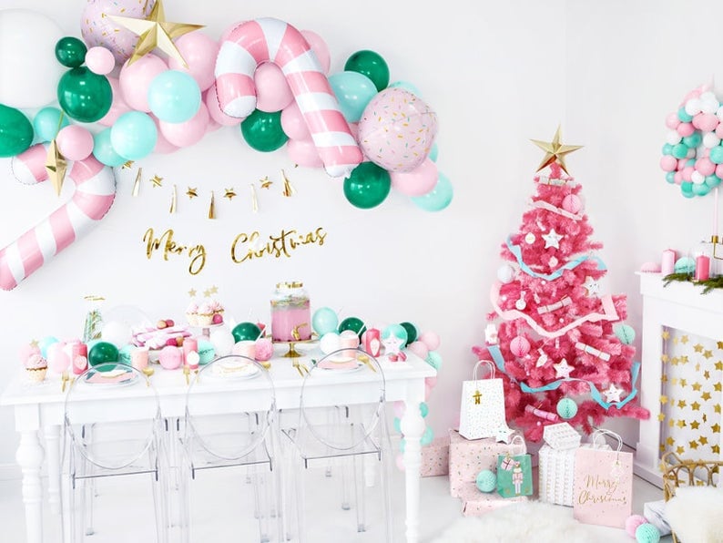 May include: A pink Christmas tree decorated with white and blue ribbons and ornaments. The tree is surrounded by white gift bags with gold stars and a sign that says "Merry Christmas". There are also pink and green balloons in the background.