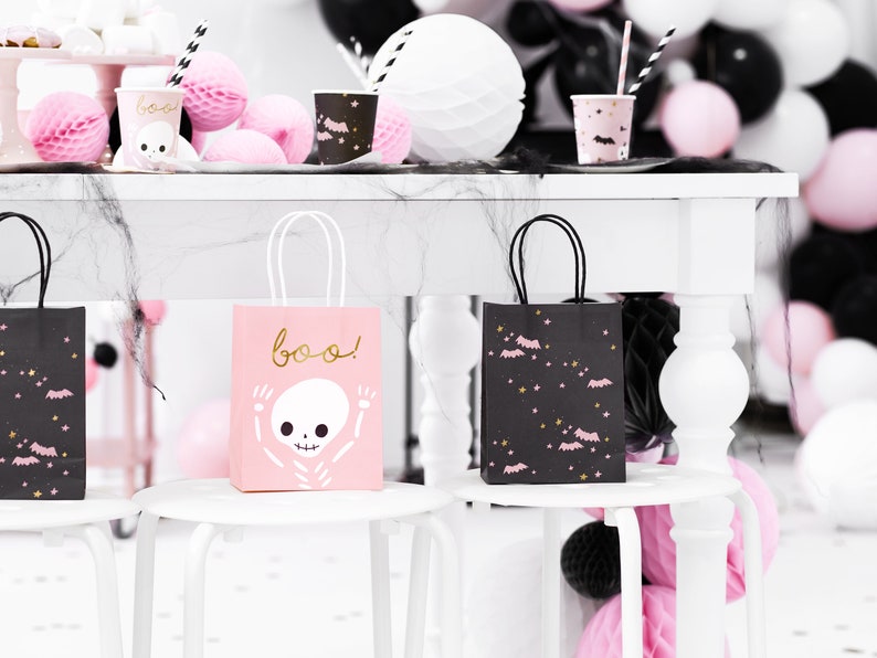 K&ouml;nnte beinhalten: Zwei Halloween-Geschenkbeutel, einer rosa mit einem Skelett und dem Text "boo!" und der andere schwarz mit rosa Flederm&auml;usen und Sternen. Die Beutel stehen auf wei&szlig;en Hockern mit einem wei&szlig;en Tisch im Hintergrund.