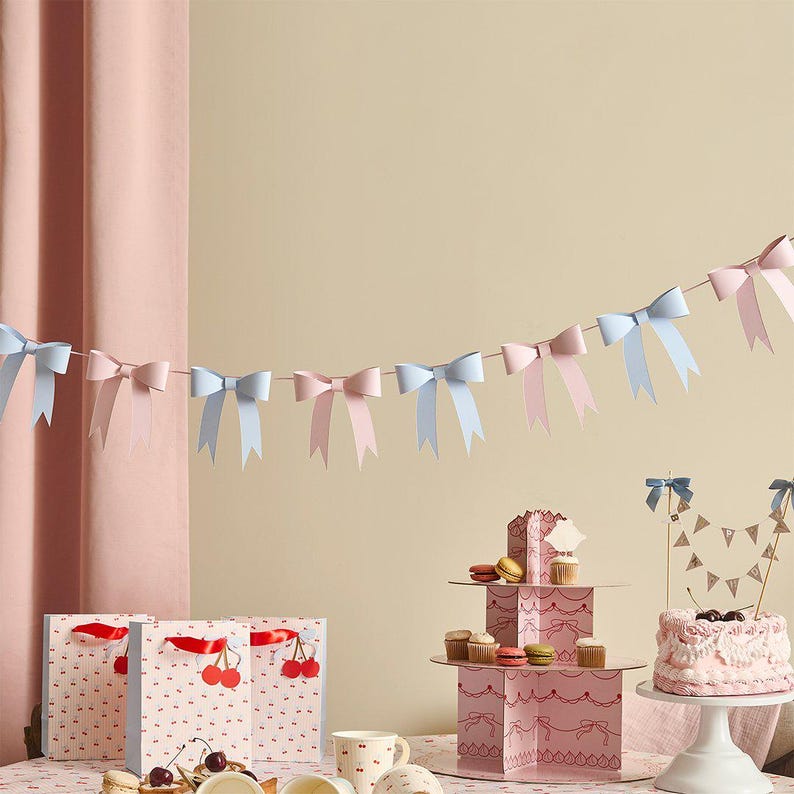 May include: A decorative garland featuring alternating light blue and pale pink paper bows strung on a thin cord. Below, three gift bags with a red cherry pattern, a tiered cardboard dessert stand with cupcakes, and a frosted cake with a banner.