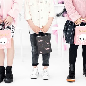 K&ouml;nnte beinhalten: Drei Kinder halten rosa und schwarze Halloween-S&uuml;&szlig;igkeitent&uuml;ten mit einem Skelett- und Fledermausdesign. Die Taschen haben den Schriftzug "Boo!"