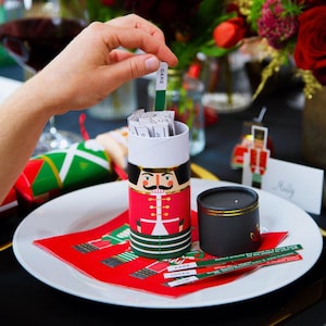 May include: A festive Christmas game set featuring a nutcracker design. The cylindrical container is red with a nutcracker illustration and holds small cards. A hand pulls a green card labeled "DARE". The set includes a small black candle holder and a red napkin.