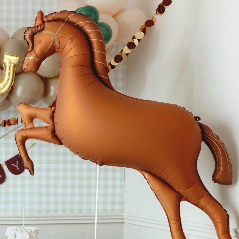 以下が含まれることがあります： メタリック仕上げの大きな茶色の馬型バルーンで、跳躍中です。バルーンには、たてがみ、尾、手綱などの詳細な特徴があります。他のバルーンやバナーとともに、パーティーの装飾の一部です。