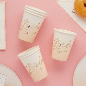 May include: Three white paper cups with the text "Bride to be" in gold script. The cups have a gradient color from white to peach. The cups are on a pink surface.