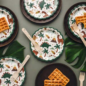 May include: A top-down view of a party table setting with dinosaur-themed paper plates, wooden forks, and waffles. The plates have a white background with a green scalloped edge and feature illustrations of dinosaurs, palm trees, and volcanoes. The plates are on dark brown plates.