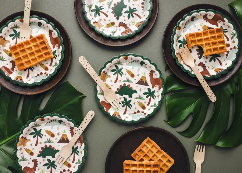 May include: A top-down view of a party table setting with dinosaur-themed paper plates, wooden forks, and waffles. The plates have a white background with a green scalloped edge and feature illustrations of dinosaurs, palm trees, and volcanoes. The plates are on dark brown plates.