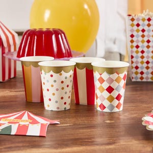 Puede incluir: Cuatro vasos de papel con bordes dorados festoneados. Las tazas presentan diferentes diseños: lunares, rayas y un patrón de diamantes. Un postre de gelatina roja, un globo amarillo y una tarjeta con temática de circo están en el fondo.
