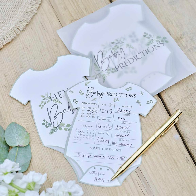 May include: White baby prediction cards shaped like onesies, with green botanical accents and the text "Baby Predictions." One card is filled out with baby details, including the name "Harry," and measurements. A gold pen rests on the wooden surface.