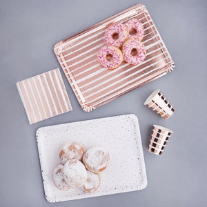 Op de afbeelding: Een set roze en witte gestreepte papieren borden en servetten met een roosgoud folie ontwerp. De borden en servetten zijn gerangschikt op een grijs oppervlak met donuts en bekers.