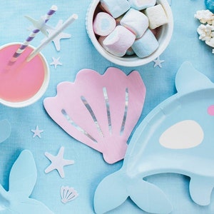 Pode incluir: Uma mesa de festa com tema de sereia com uma concha rosa, pratos de golfinho azuis e limonada rosa em um copo transparente com dois canudos listrados.