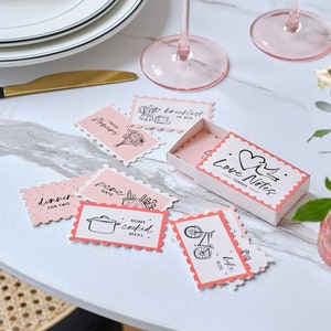 May include: A pink and white matchbox with the text "Love Notes - Tokens" on the front. The matchbox contains small pink and white cards with various activities written on them, such as "Dinner for Two", "Picnic Date", "Home Cooked Meal", "Bike Ride", "Fresh Flowers", and "Breakfast in Bed".